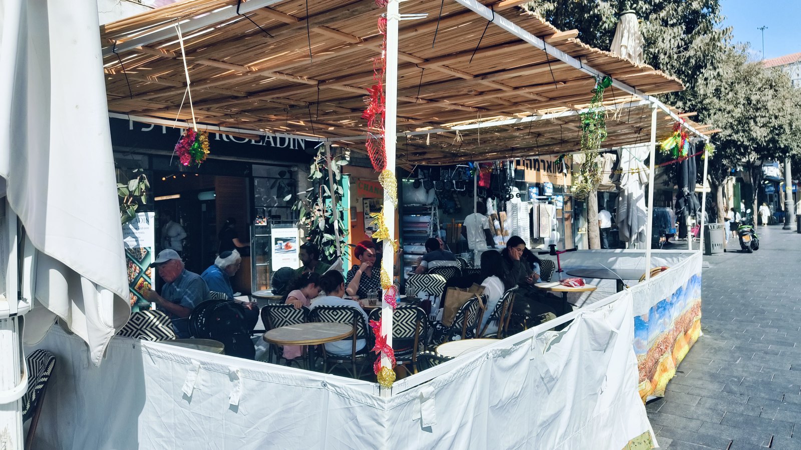 A sukkah with a roof of branches set up outside a cafe on a Jerusalem street, people eating at tables inside it